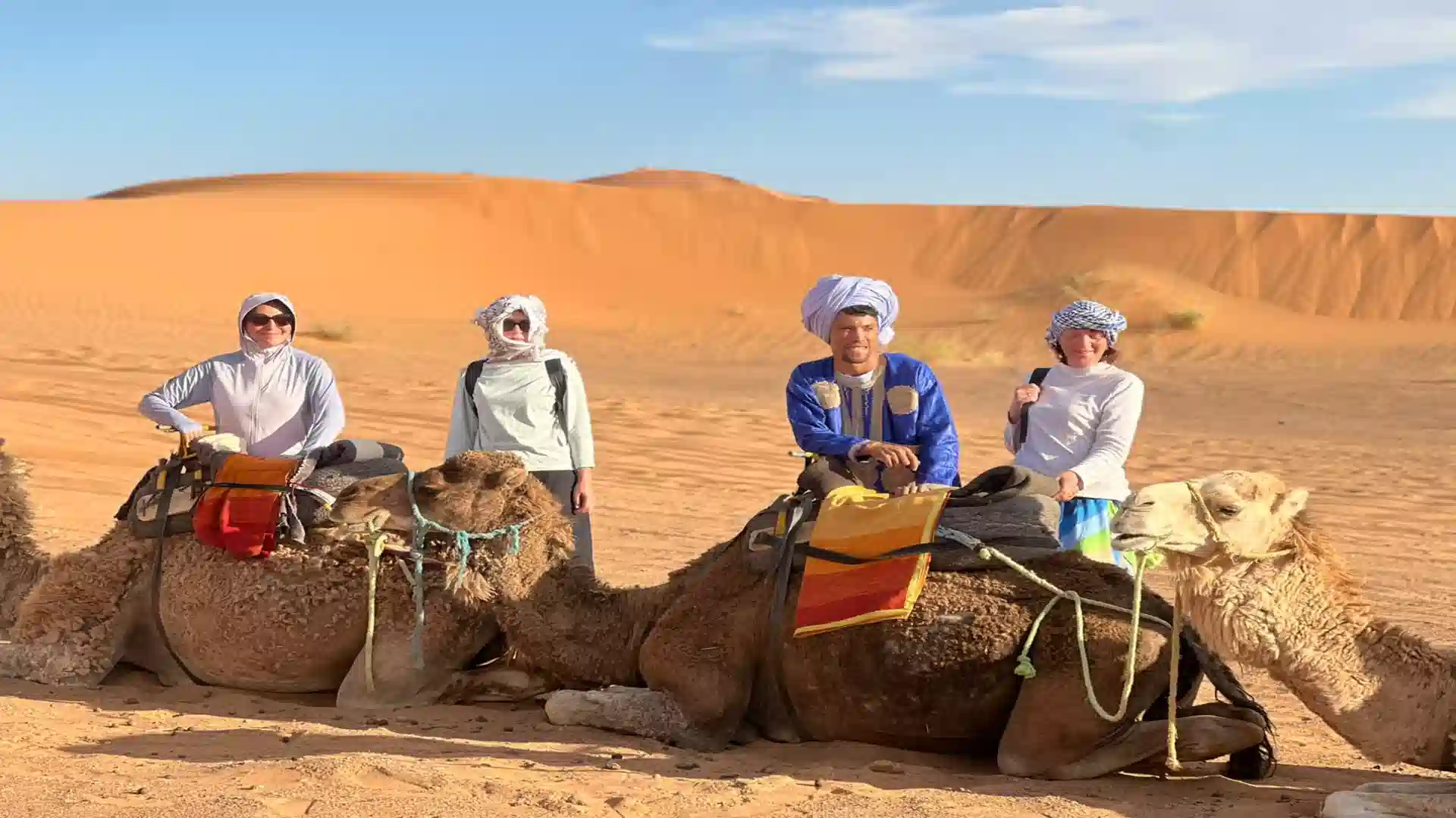 Travelers wearing traditional headscarves posing with camels and a local guide in the golden sand dunes of Merzouga Sahara Desert. 3 days tour from Fes to Marrakech