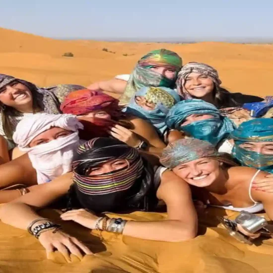 Group of happy travelers lying together on the golden sand dunes of the Merzouga Sahara Desert wearing colorful headscarves.5 days tour from Marrakech to Merzouga- Over Morocco Tours