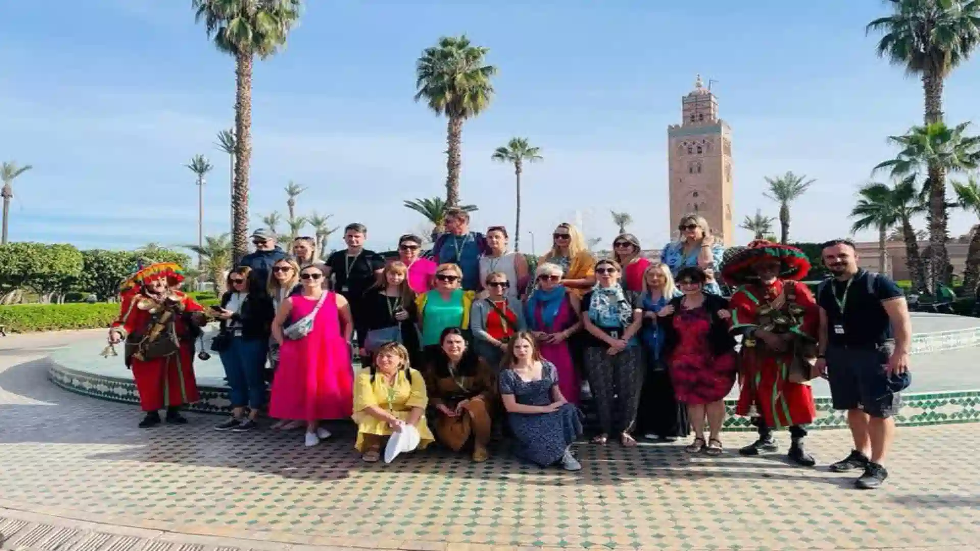 A group of tourists posing for a photo in front of the Koutoubia Mosque minaret and palm trees in Marrakech, accompanied by traditional water sellers. things to do in Marrakech