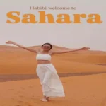 A woman in a white outfit standing on golden sand dunes with the text Habibi welcome to Sahara on a Morocco in December