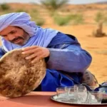 A man wearing a light blue turban and blue robe dusts sand from a large, circular Moroccan flatbread (khobz) that was baked in the Sahara desert sand.