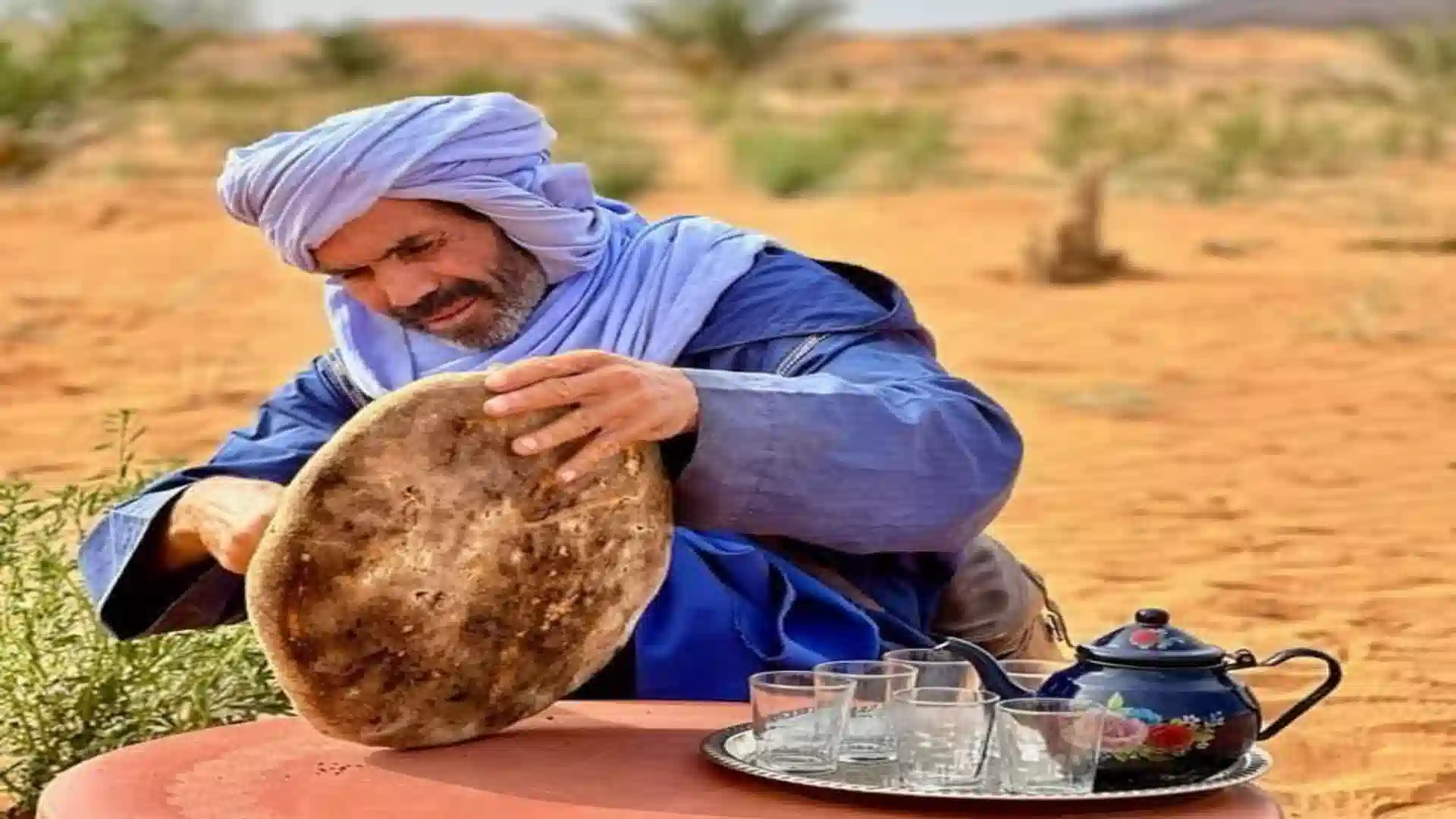 A man wearing a light blue turban and blue robe dusts sand from a large, circular Moroccan flatbread (khobz) that was baked in the Sahara desert sand.