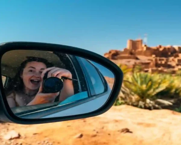 Side mirror reflection of a smiling woman taking a photo with a camera during a road trip in Morocco, with a traditional clay kasbah village blurred in the background.